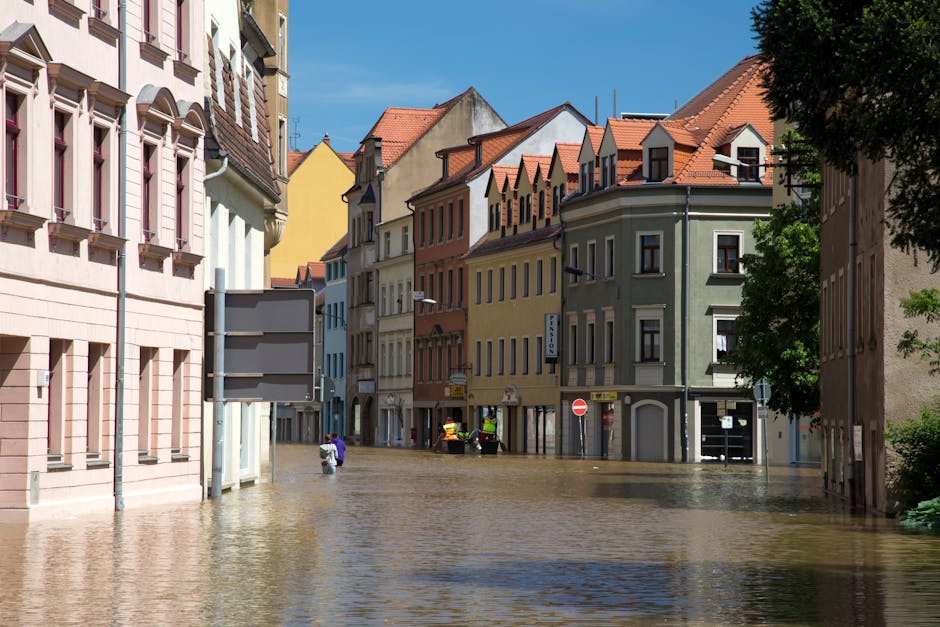 A street scene in Arnos Grove showing multiple multi-storey buildings with pastel-colored facades, including pink, yellow, and green, under bright daylight. The area appears flooded with water coverin