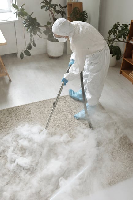 A professional cleaner dressed in a full white protective suit, including gloves, a mask, and boot covers, is performing deep carpet cleaning using a large vacuum machine with a hose. The scene takes 